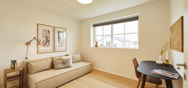 Fleksibelt værelse fra eurodan-huse med beige daybed, lille sidebord, sort skrivebord, rosa stol, væglampe, to indrammede plakater og bredt vindue – inspiration til kontor, gæsteværelse og børneværelse i nybyggeri.