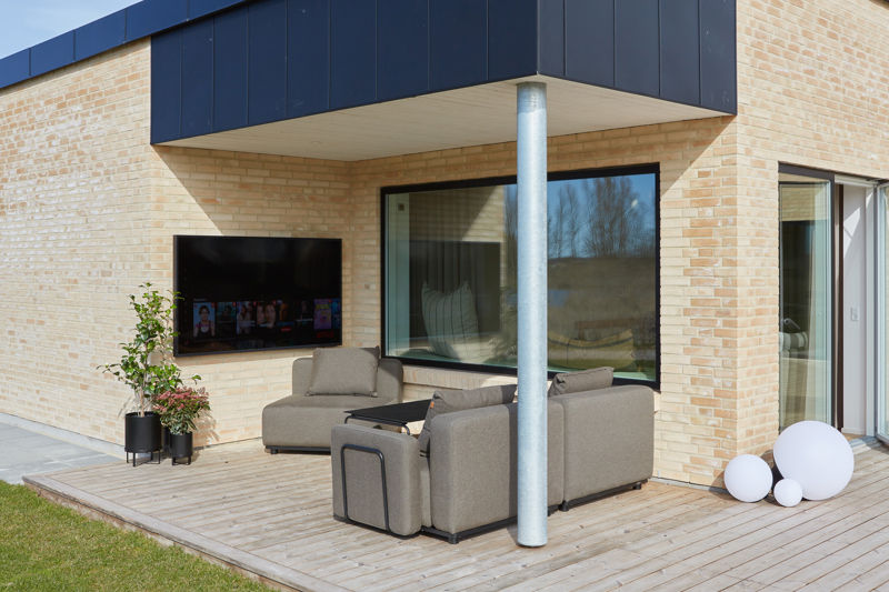 Overdækket træterrasse fra eurodan-huse med sort udhæng, synlig stålsøjle, stor udendørs sofa, vægmonteret tv, plantekrukker og store vinduespartier mod opholdsrummet – inspiration til nybyggeri med udekøkkenstemning, loungeområde og husdrømme.