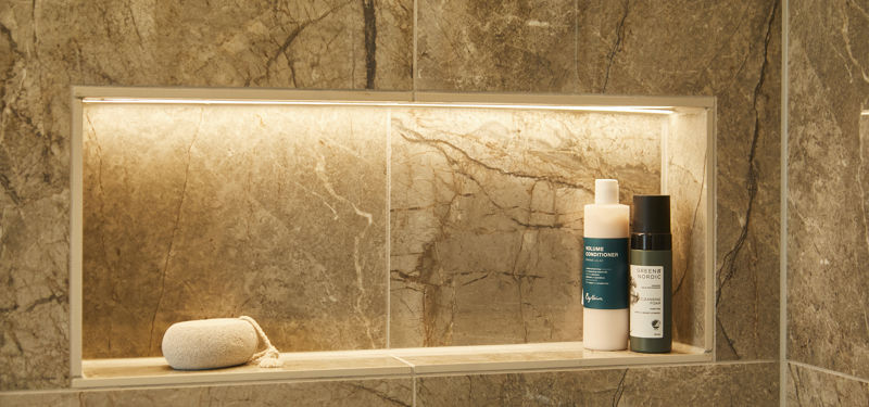 Detalje fra badeværelse fra eurodan-huse med indbygget oplyst vægniche i marmorerede fliser, hylde til shampoo og sæbe, vaskeklud og varme stenfarver – inspiration til gennemtænkte badeværelsesdetaljer i nybyggeri, husbyggeri og moderne husdrømme.