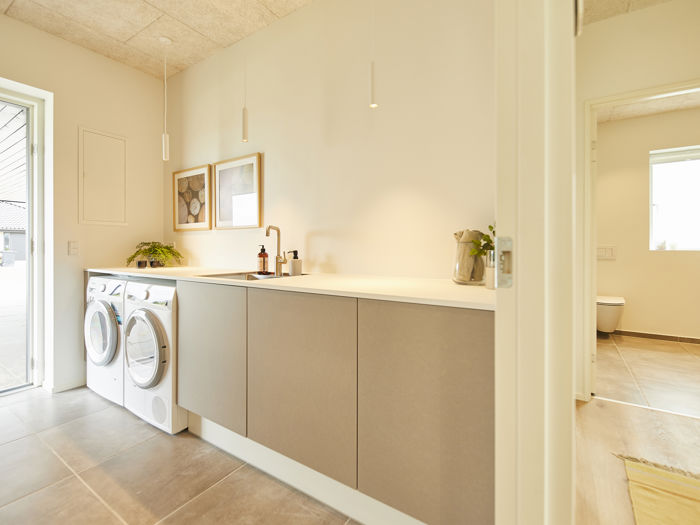 Lyst bryggers fra eurodan-huse med vaskemaskine og tørretumbler ved terrassedør, lang hvid bordplade, beige underskabe, sort armatur, vægbilleder og adgang til badeværelse i højre side – inspiration til nybyggeri med funktionel indretning og praktiske husdrømme.