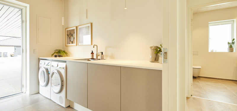 Lyst bryggers fra eurodan-huse med vaskemaskine og tørretumbler ved terrassedør, lang hvid bordplade, beige underskabe, sort armatur, vægbilleder og adgang til badeværelse i højre side – inspiration til nybyggeri med funktionel indretning og praktiske husdrømme.
