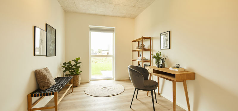 Lyst hjemmekontor med træskrivebord, polstret stol, åben reol, bænk, grønne planter og stort vindue – funktionel inspiration til moderne nybyg og husdrømme.