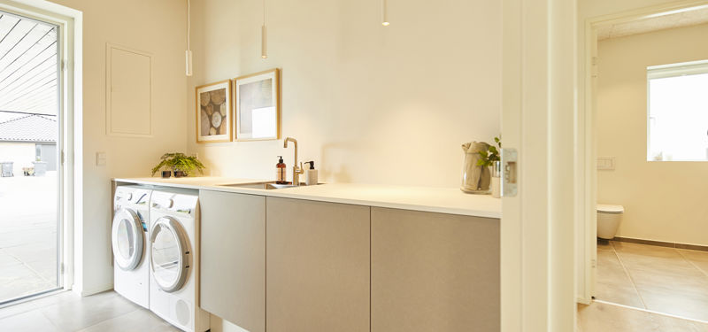 Lyst bryggers med vaskemaskine, tørretumbler, lang skabsløsning med bordplade og vask– praktisk inspiration til moderne nybyg og husdrømme.