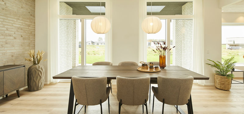 Lys spiseplads med træbord, polstrede stole, to pendellamper, høje vinduespartier, lyst trægulv og hvid murstensvæg – indbydende inspiration til moderne nybyg og husdrømme.