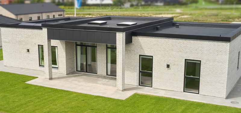 Moderne H-hus med lys murstensfacade, fladt sort tag, høje vinduespartier og overdækket terrasse ud mod en grøn have – stilren inspiration til nybyg og husdrømme.