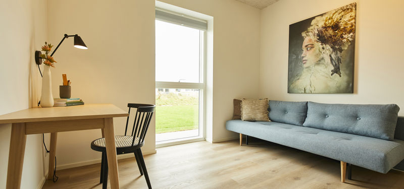 Lyst gæsteværelse med blå sovesofa, skrivebord i træ, sort stol, stort vindue og moderne vægkunst – fleksibel inspiration til nybyg og familiens husdrømme.