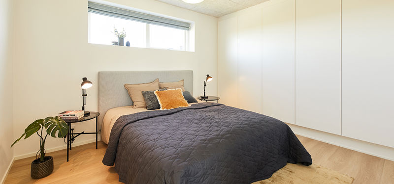 Soveværelse fra eurodan-huse med dobbeltseng, mørk quiltet sengetæppe, polstret sengegavl, sort sengeborde, små læselamper, langt vandret vindue og stor hvid skabsvæg fra gulv til loft – inspiration til funktionel opbevaring i nybyggeri og moderne husdrømme.