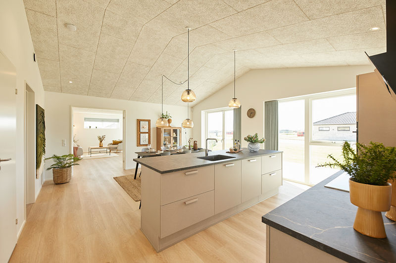 Åbent køkken-alrum fra eurodan-huse med lys køkkenø, mørk bordplade, sort vask, tre hængende glaskupler over arbejdszonen, store vinduer mod terrassen og kig videre til spiseplads og stue – inspiration til nybyggeri med god sammenhæng mellem køkken og opholdsrum.