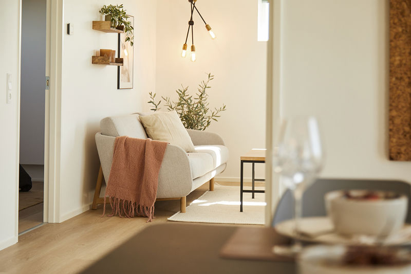 Nært kig til stue fra eurodan-huse med lys lænestol, rosa plaid, lille sort sidebord, væghylder med planter, pendellampe med synlige pærer og sløret spisebord i forgrunden – inspiration til nybyggeri, husbyggeri og hyggelig stueindretning.