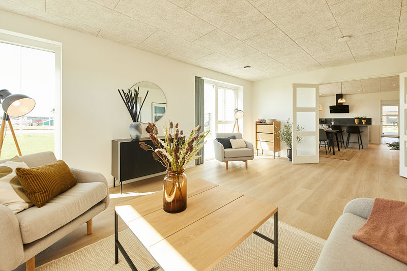 Rummelig stue fra eurodan-huse med lyse sofaer, stort sofabord i træ, brun vase med tørrede blomster, sort skænk med rundt spejl, lænestol ved vinduet og kig til køkken-alrum – inspiration til nybyggeri, husbyggeri og åbne husdrømme.