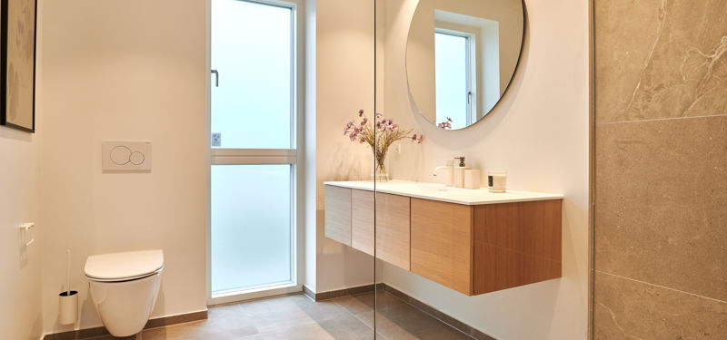 Lyst og moderne badeværelse i nybyggeri fra eurodan-huse med væghængt toilet, rummeligt badeværelsesmøbel i træ, rundt spejl og store beige fliser, der skaber en eksklusiv og rolig indretning til moderne husdrømme.
