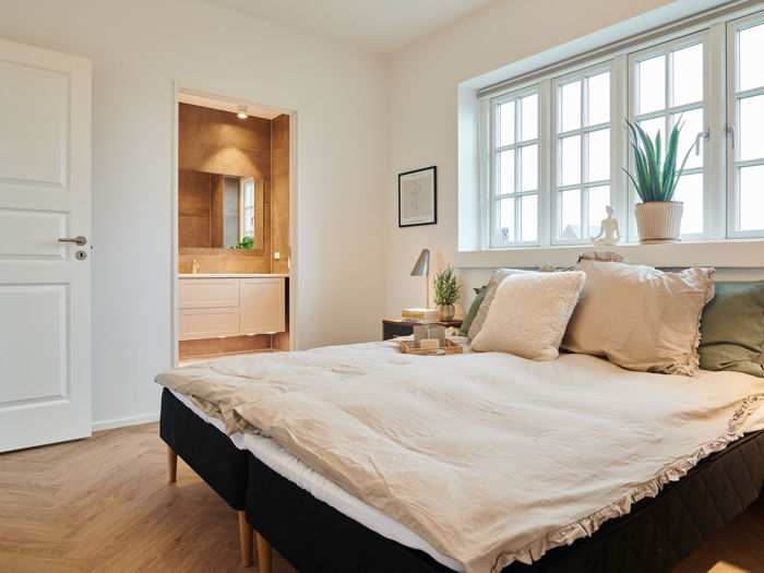 Soveværelse fra eurodan-huse med sprossede vinduer over sengen, bred vindueskarm med plante, beige sengetøj, mørke sengeborde og kig til badeværelse gennem åben dør – inspiration til nybyggeri, husbyggeri og rolige rum med naturligt lys.