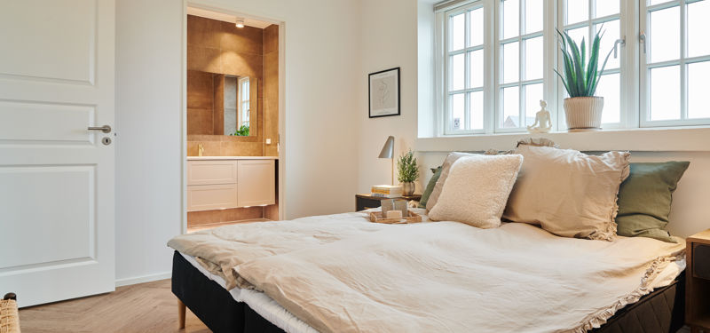 Soveværelse fra eurodan-huse med sprossede vinduer over sengen, bred vindueskarm med plante, beige sengetøj, mørke sengeborde og kig til badeværelse gennem åben dør – inspiration til nybyggeri, husbyggeri og rolige rum med naturligt lys.