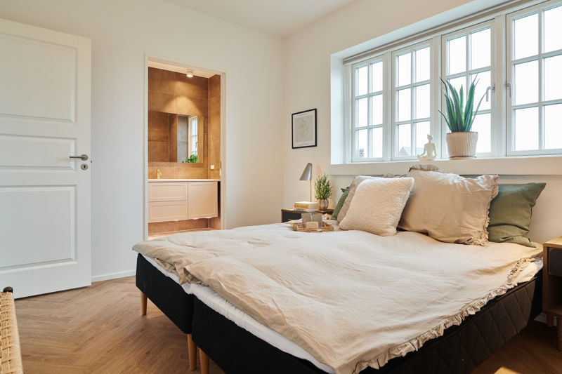 Soveværelse fra eurodan-huse med sprossede vinduer over sengen, bred vindueskarm med plante, beige sengetøj, mørke sengeborde og kig til badeværelse gennem åben dør – inspiration til nybyggeri, husbyggeri og rolige rum med naturligt lys.