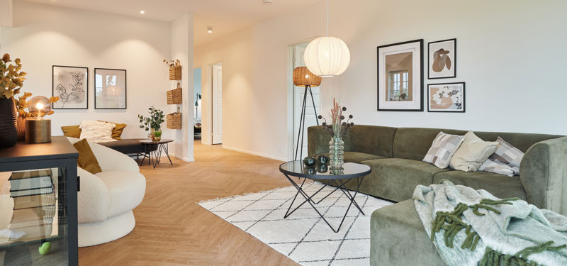 Stue fra eurodan-huse med grøn hjørnesofa, cremefarvet loungestol, sort rundt sofabord, hvidt tæppe med grafisk mønster, billedvæg over sofaen og varm belysning i rummet – inspiration til nybyggeri, husbyggeri og moderne husdrømme.