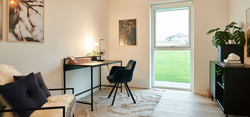 Hjemmekontor fra eurodan-huse med smalt skrivebord, mørk kontorstol, væglampe, loungestol med skind, sort reol, stor plante og højt vindue med udsyn til græs – inspiration til kontor i nybyggeri og moderne husdrømme.