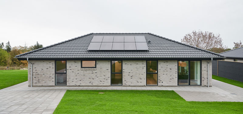 Længehus i nybyggeri med lys murstensfacade, sort valmtag, solceller og store vinduespartier – inspiration til moderne husdrømme.