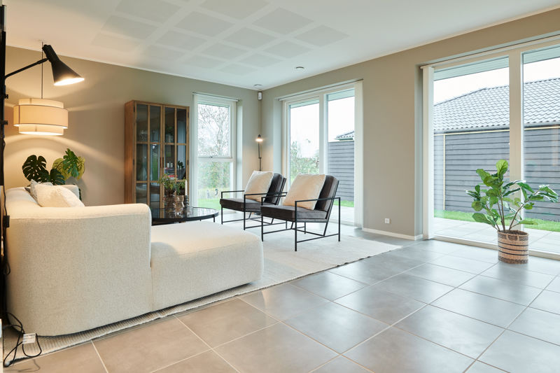 Stue fra eurodan-huse med lys sofa, to sorte loungestole, rundt glasbord, vitrineskab i træ, store vinduer mod terrassen, grøn plante i kurv og mørke klinker på gulvet – inspiration til nybyggeri, husbyggeri og funktionelle opholdsrum.