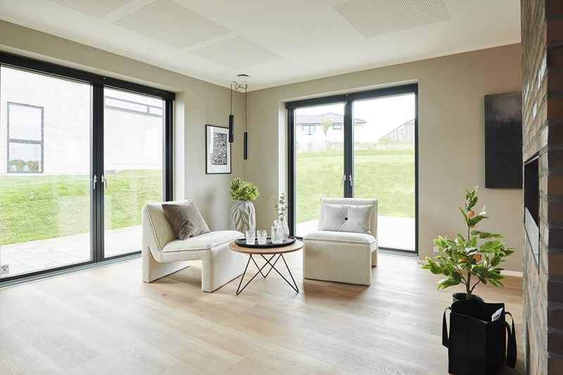 Lys stue fra eurodan-huse med to store sorte terrassedøre, brede vinduespartier, lyse loungestole, rundt sofabord, plante ved pejs og akustikloft – inspiration til nybyggeri, husbyggeri og opholdsrum med udsyn til haven.