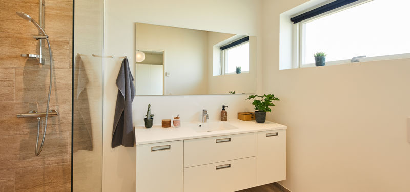 Badeværelse fra eurodan-huse med hvidt badmøbel, bred spejlvæg, højtsiddende vindue med sort rullegardin, grønne planter på bordpladen og bruseniche med trælook fliser og glasvæg – inspiration til lyst nybyggeri, husbyggeri og praktiske husdrømme.