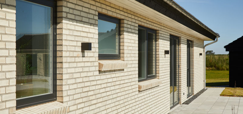 Facade på moderne længehus i nybyggeri med lyse mursten, sorte vinduesrammer og flisebelagt gangsti – inspiration til stilrent husbyggeri.