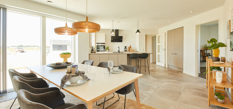 Rummeligt køkken-alrum med beige køkken, stort spisebord, fletpendler, barstole og glasdøre mod terrassen – hjemmets naturlige samlingspunkt.