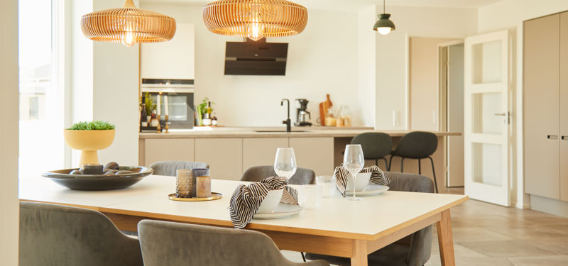Dækket spisebord med polstrede stole og fletpendler foran et moderne beige køkken med sorte detaljer og siddepladser ved køkkenbordet.