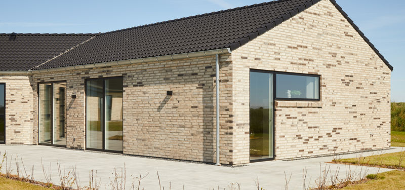 Moderne knækhus med flisebelagt terrasse, lyse mursten, sorte vinduespartier og mørkt tegltag – inspiration til uderummet omkring et nyt hus.