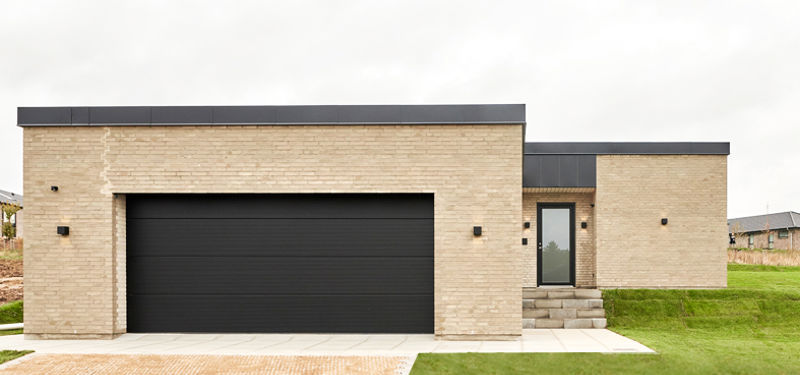 Facade med lys mursten, sort dobbelt garageport, fladt tag, trin op til indgangsdør og bred flisebelagt indkørsel foran garagen – inspiration til integreret garage i nybyggeri og praktiske husdrømme fra eurodan-huse.