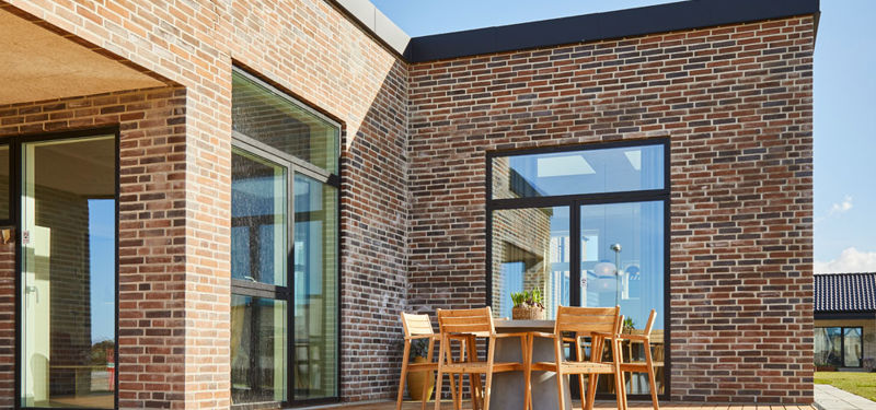 Solrig træterrasse ved moderne nybyggeri med rød murstensfacade, fladt tag, store sorte vinduespartier og spisebord med havestole – inspiration til udeliv og husdrømme.