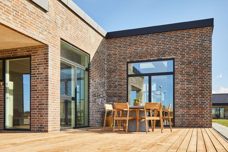 Solrig træterrasse ved moderne nybyggeri med rød murstensfacade, fladt tag, store sorte vinduespartier og spisebord med havestole – inspiration til udeliv og husdrømme.
