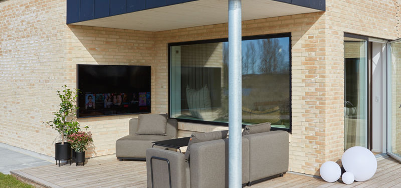 Overdækket træterrasse ved et moderne murstenshus med loungeområde, væghængt TV og store vinduespartier – inspiration til udeliv i et nyt eurodan-hus.
