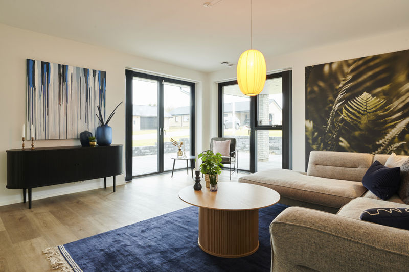 Stue fra eurodan-huse med sorte vinduesrammer, hjørnevinduer, beige hjørnesofa, rundt sofabord, blå tæppe, sort skænk og stor pendellampe – inspiration til nybyggeri, husbyggeri og moderne stue med godt lysindfald.