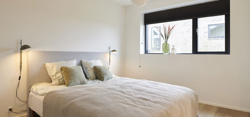 Lyst soveværelse fra eurodan-huse med dobbeltseng, lyse puder, beige sengetæppe, stofbeklædt sengegavl, sorte væglamper og bredt sort vindue med plante i vindueskarmen – inspiration til rolig soveværelsesindretning i nybyggeri og nye husdrømme.