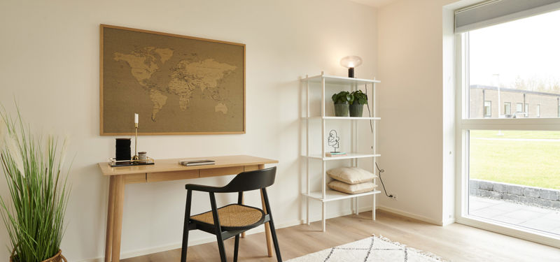 Lyst kontor fra eurodan-huse med træskrivebord, sort stol med fletsæde, vægkort, hvid reol, grøn plante og stor terrassedør med dagslys – inspiration til hjemmearbejdsplads i nybyggeri og gennemtænkt husbyggeri.