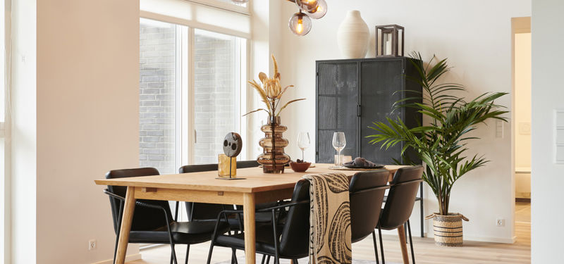 Moderne spiseplads med træbord, sorte stole, skulpturel pendel, høje vinduespartier, mørkt vitrineskab og grønne planter – elegant inspiration til nybyg, drømmehuset og familiens husdrømme.