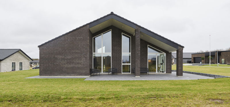 Moderne vinkelhus i nybyggeri med mørk murstensfacade, markant saddeltag og store gulv-til-loft-vinduer – inspiration til arkitektoniske husdrømme.