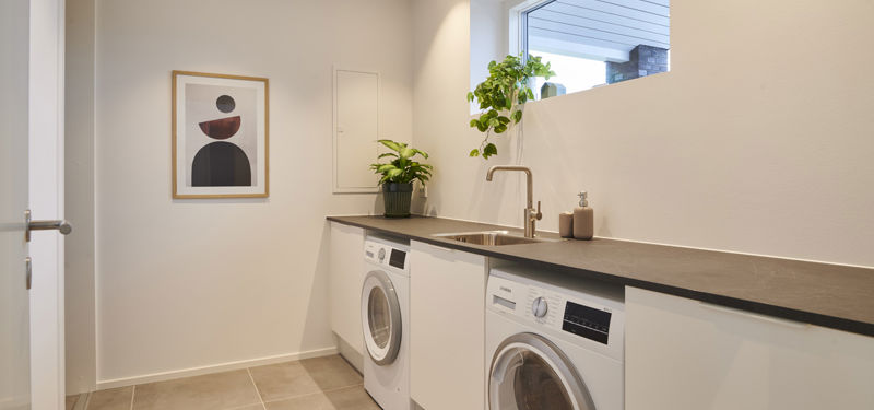 Bryggers fra eurodan-huse med hvid vaskemaskine og tørretumbler under mørk bordplade, nedfældet vask, højt smalt vindue med grøn plante i karmen og lys væg med grafisk plakat – inspiration til praktisk nybyggeri, funktionelt husbyggeri og gennemtænkte husdrømme.
