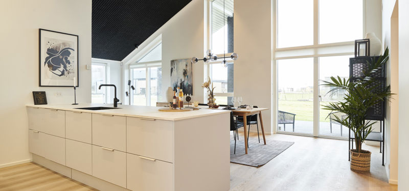 Lyst køkken-alrum med hvid køkkenø, højt skråt loft med mørk træbeklædning, store vinduespartier, spiseplads og lyst trægulv – moderne inspiration til nybyg og husdrømme.