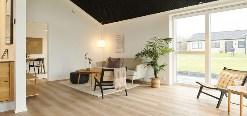 Lyst opholdsrum med loungestole, bænk, grøn plante, lyst trægulv, mørkt loft og stort vinduesparti mod haven – moderne inspiration til nybyg og husdrømme.
