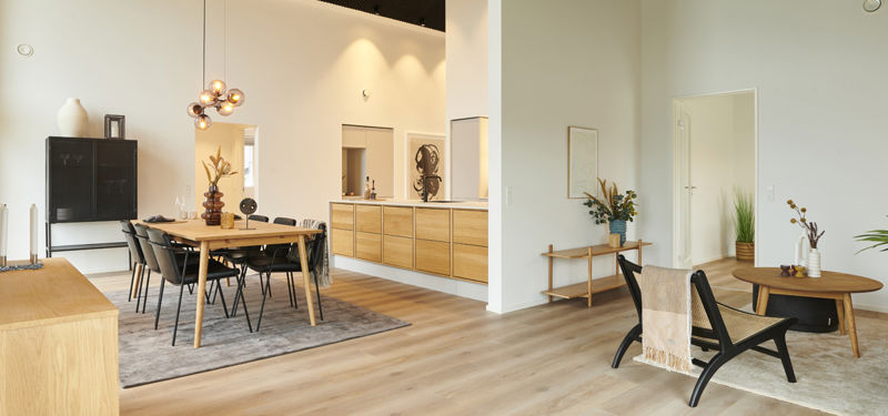 Rummeligt køkken-alrum med spiseplads, lyst trægulv, højt mørkt listeloft, køkken i lyst træ og hyggeligt opholdsrum med loungestole – moderne inspiration til nybyg og husdrømme.