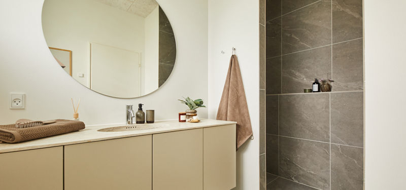 Badeværelse fra eurodan-huse med langt beige badmøbel, rund spejlflade, hvid bordplade, håndklæde på væggen og bruseniche med store mørke fliser og indbygget hylde – inspiration til funktionelt nybyggeri, husbyggeri og moderne badeværelsesdrømme.