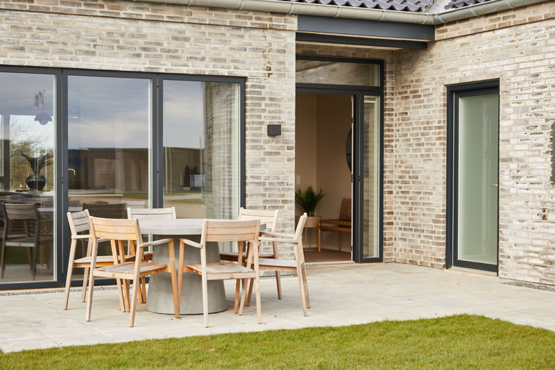 Terrasse ved facade fra eurodan-huse med store skydedørspartier, mørke rammer, åben hoveddør med sidevindue, smal høj dør og spiseplads med træmøbler udenfor – inspiration til vinduer, døre og uderum i nybyggeri.
