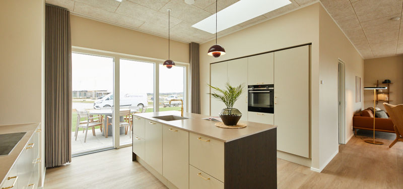 Lyst køkken fra eurodan-huse med køkkenø i beige og mørkt træ, messinggreb, indbygget ovn i høj skabsvæg, to røde pendler, ovenlys, store vinduer med adgang til terrasse og kig til stueområde med varm lænestol – inspiration til åbent køkken-alrum i nybyggeri og nye husdrømme.