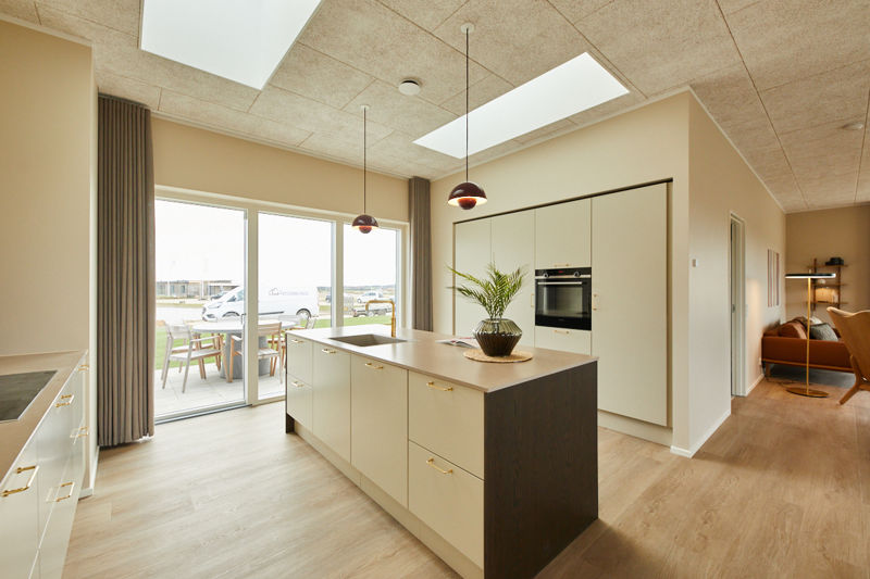 Lyst køkken fra eurodan-huse med køkkenø i beige og mørkt træ, messinggreb, indbygget ovn i høj skabsvæg, to røde pendler, ovenlys, store vinduer med adgang til terrasse og kig til stueområde med varm lænestol – inspiration til åbent køkken-alrum i nybyggeri og nye husdrømme.