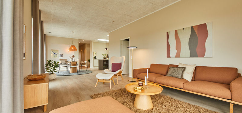 Lang stue fra eurodan-huse med cognacfarvet sofa, hvid loungestol med fodskammel, rundt træbord, fluffy tæppe, store gardiner og kig til spiseplads i baggrunden – inspiration til nybyggeri, husbyggeri og åbne opholdsrum.