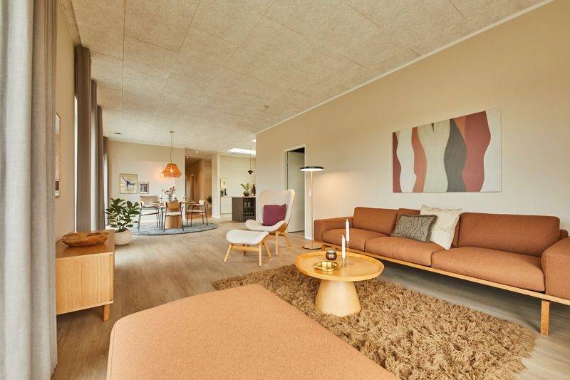 Lang stue fra eurodan-huse med cognacfarvet sofa, hvid loungestol med fodskammel, rundt træbord, fluffy tæppe, store gardiner og kig til spiseplads i baggrunden – inspiration til nybyggeri, husbyggeri og åbne opholdsrum.