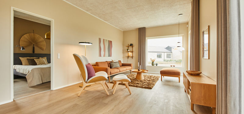 Stue fra eurodan-huse med cognacfarvet sofa, hvid loungestol, fodskammel, rundt træbord, langt gardinparti, træskænk og kig til soveværelse gennem åben dør – inspiration til nybyggeri, husbyggeri og velindrettede husdrømme.