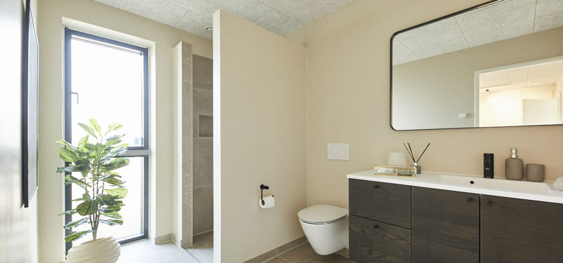 Moderne badeværelse med mørkt badmøbel, hvid bordplade, stort spejl, væghængt toilet, bruseniche, højt vindue og grøn plante – stilren inspiration til nybyg og husdrømme.