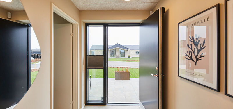 Entré fra eurodan-huse med åben sort hoveddør, stort vinduesparti, lyse gulvfliser, rundt spejl, vægplakater og polstret bænk i hjørnet – inspiration til indbydende nybyggeri, entrédesign og moderne husdrømme.